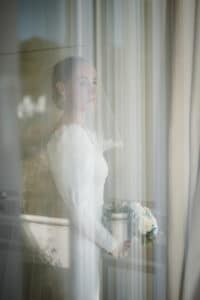 Eine Braut steht an einem Fenster. Sie trägt ein langärmeliges weißes Brautkleid und einen Schleier. Sie hält einen Strauß weißer Blumen in der Hand und blickt hinaus. Weiches, natürliches Licht beleuchtet ihre Figur und fängt die heitere Vorfreude auf ihre Hochzeit ein.