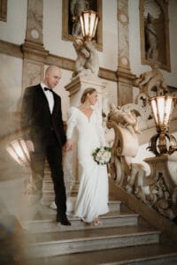 Braut und Bräutigam steigen anmutig Hand in Hand eine verzierte Treppe hinab. Die Braut, prächtig in ihrem eleganten weißen Kleid und Schleier, hält einen Blumenstrauß, während der Bräutigam in einem schwarzen Smoking glänzt. Die Treppe, ideal für eine romantische Hochzeit, ist mit Vintage-Lampen und Skulpturen geschmückt.