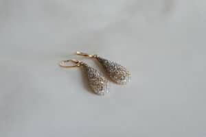 Ein Paar elegante, tropfenförmige Ohrringe mit aufwendigen Details, perfekt für eine Hochzeit, verfügen über einen Farbverlauf aus kleinen funkelnden Steinen und sind nebeneinander auf einer weichen, weißen Oberfläche platziert.