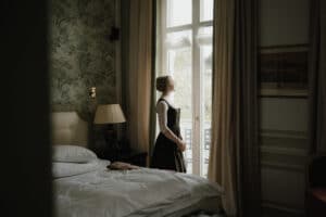 Eine Frau in einem langen Kleid steht in einem schwach beleuchteten Raum am Fenster und stellt sich ihre perfekte Hochzeit vor. Sie blickt nach draußen, umgeben von elegantem Dekor und einem ordentlich gemachten Bett. Sanftes Tageslicht fällt durch die transparenten Vorhänge und bereitet die Bühne für ihren Traum vom Heiraten.