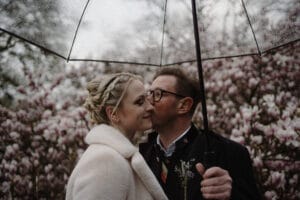 Ein Paar steht unter einem durchsichtigen Schirm, umgeben von blühenden rosa Blumen. Der Mann küsst die Frau auf die Wange, beide sind in warme Kleidung gehüllt. Die Atmosphäre ist romantisch und heiter – ein bezaubernder Moment, der Träume von Hochzeitsglück weckt.
