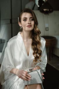Eine Frau mit langem, welligem Haar sitzt in einem sanft beleuchteten Raum und trägt ein weißes Satingewand. Während sie sich auf ihre Hochzeit vorbereitet, hält sie eine silberne, strukturierte Clutch in den Händen. Ihr ruhiger und nachdenklicher Gesichtsausdruck verrät die Bedeutung dieses Moments in ihrer Hochzeitsplanung.