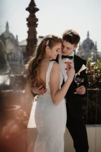 Eine Braut im Spitzenkleid und ein Bräutigam im Smoking umarmen sich liebevoll auf einem Dach und feiern ihre Hochzeit. Sie halten Champagnergläser in den Händen, umgeben von kunstvoller Architektur und Grün unter dem klaren Himmel.
