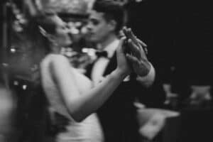 Ein Schwarzweißfoto zeigt ein eng tanzendes Paar, dessen verschränkte Hände Liebe und Heirat symbolisieren. Ihre Gesichter und Körper sind sanft verschwommen, wodurch eine verträumte, intime Atmosphäre entsteht, die an die Romantik des Hochzeitstags erinnert. Der schwach beleuchtete Hintergrund trägt zur bezaubernden Stimmung bei.
