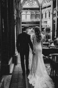 Ein Schwarzweißfoto eines Paares, das Händchen hält und durch eine große, reich verzierte Halle geht. Die Frau trägt ein langes, elegantes Hochzeitskleid und der Mann einen Anzug. Sie sind von hinten zu sehen, umgeben von luxuriöser Architektur – ein zeitloser Moment der Hochzeitsmagie.