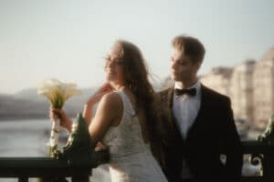 Das Paar steht, vertieft in die Hochzeitsplanung, auf einer malerischen Brücke. Die Frau im weißen Kleid strahlt, während sie einen Blumenstrauß hält, während der Mann im schwarzen Anzug sie liebevoll ansieht. Hinter ihnen liegen der Fluss und die Gebäude unter einem weichen, dunstigen Himmel.