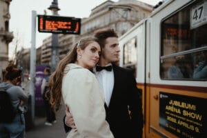 Ein Paar in formeller Kleidung, das vielleicht von seiner zukünftigen Hochzeit träumt, umarmt sich in der Nähe einer Straßenbahnhaltestelle in einer städtischen Umgebung. Die Frau trägt einen eleganten beigen Mantel und der Mann einen schwarzen Smoking. In der Nähe stehen Menschen, im Hintergrund ist eine Straßenbahn zu sehen, auf der Werbung für eine bevorstehende Show gezeigt wird.