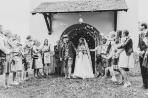Ein Schwarzweißfoto fängt eine freudige Hochzeitsszene ein. Braut und Bräutigam verlassen eine Kirche, umgeben von lächelnden Gästen. Während sie mit Blütenblättern überschüttet werden, feiern mehrere Personen, darunter ein Kind, in traditioneller Kleidung diesen schönen Moment auf der Hochzeitsreise des Paares.