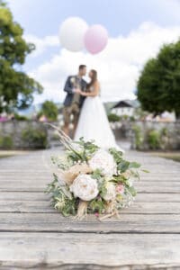 Ein Brautstrauß ruht auf den Holzbrettern einer Brücke, im Hintergrund sind Braut und Bräutigam mit Luftballons zu sehen. Die Szene strahlt den Charme einer Hochzeitsfeier im Freien aus, umgeben von Grün und einem teilweise bewölkten Himmel – eine perfekte Momentaufnahme einer durchdachten Hochzeitsplanung.