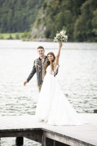 Ein fröhliches Paar steht auf einem Holzsteg an einem See und feiert seine Hochzeit. Die Braut, die ein weißes Kleid trägt, streckt lächelnd ihren Blumenstrauß in die Höhe, während der Bräutigam in einer grauen Jacke stolz neben ihr steht. Üppiges Grün bietet die perfekte Kulisse für ihren besonderen Tag.