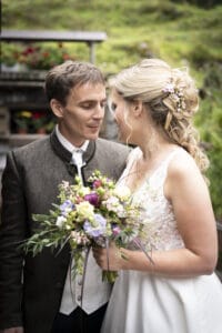 Ein Paar steht eng beieinander, lächelt sich an und verkörpert die Freude der Hochzeit. Die Frau hält einen bunten Blumenstrauß und trägt ein weißes Kleid mit Blumendetails. Ihr Partner trägt eine traditionelle Jacke über einem weißen Hemd. Sie sind draußen, umgeben von üppigem Grün, während sie ihre Hochzeit feiern.