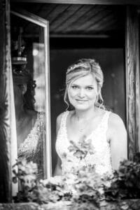 Schwarzweißfoto einer Braut mit hellem Haar und Blumenkopfschmuck, die ein Spitzenkleid trägt. Sie steht an einem Fenster und blickt in die Kamera. Blumen zieren den Vordergrund und fangen einen eleganten Moment ihrer Hochzeit ein.