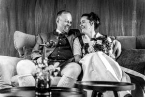 Ein Schwarzweißfoto zeigt ein Paar, das in die Hochzeitsplanung vertieft ist. Es ist in traditioneller Kleidung gekleidet, sitzt auf einem Sofa und lächelt sich an. Der Mann trägt eine Jacke und Kniestrümpfe, während die Frau einen Blumenstrauß hält. Sie teilen einen freudigen, intimen Moment.
