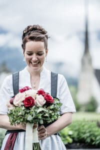 Eine lächelnde Frau in traditioneller Kleidung hält einen bunten Rosenstrauß in der Hand und verkörpert damit die Freude einer Hochzeit. Im Hintergrund sind üppiges Grün und ein verschwommener Kirchturm zu sehen – perfekt für alle, die von einer bezaubernden Hochzeit unter bewölktem Himmel träumen.