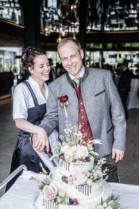 Ein lächelndes Paar in traditioneller Kleidung schneidet eine weiße, mit Blumenarrangements und Beeren geschmückte Torte an und feiert seine Hochzeit. Sie befinden sich in einem Raum mit reflektierender Decke und durch die Fenster ist eine üppige grüne Landschaft zu sehen, die ihre gut ausgeführte Hochzeitsplanung perfekt widerspiegelt.