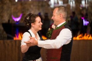Ein lächelndes Paar tanzt zusammen in traditioneller Kleidung bei seiner Hochzeit. Die Frau trägt ein schwarz-weißes Kleid, während der Mann eine rote Weste und ein weißes Hemd trägt. Sie wirken fröhlich, mit einem verschwommenen, warm beleuchteten Hintergrund, der die Essenz ihres besonderen Tages einfängt.
