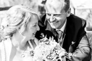 Ein Schwarzweißfoto zeigt ein lächelndes Paar bei seiner Hochzeit. Die Braut hält einen Blumenstrauß und trägt ein Kleid mit Schleier, während der Bräutigam einen Anzug trägt. Sie schauen sich liebevoll an.