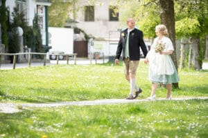 Ein Paar in traditioneller Kleidung geht Hand in Hand auf einem von Bäumen gesäumten Grasweg. Die Braut hält einen Blumenstrauß und beide lächeln, während ihre Hochzeit an einem hellen, sonnigen Tag stattfindet.