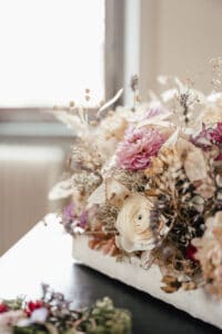 Ein Strauß Trockenblumen für die Hochzeitsplanung besteht aus pastellrosa und weißen Blüten auf einer dunklen Oberfläche. Das Arrangement besteht aus zarten Blättern und kleineren Blüten, während im Hintergrund Sonnenlicht durch ein Fenster fällt und eine sanfte, ruhige Atmosphäre schafft, die perfekt für die Planung Ihres besonderen Tages ist.