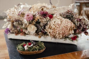 Ein dekoratives Arrangement für eine Hochzeit mit getrockneten Blumen wie Hortensien und Rosen in einem rechteckigen Gefäß. Eine kleinere Schale mit grünem Moos und kleinen Blumen steht davor. Das Ganze steht auf einem Holztisch mit einer dunklen Unterlage darunter – perfekt für eine elegante Hochzeitsplanung.