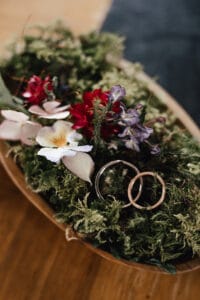 Eine Holzschale mit üppigem grünem Moos und leuchtenden Blumen – roten, violetten und weißen Blüten – fängt die Essenz einer Hochzeit perfekt ein. Zwei goldene Ringe liegen eingebettet zwischen dem Laub, elegant auf einem Holztisch platziert, und verleihen Ihrer Hochzeitsplanungsdekoration eine exquisite Note.