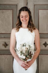 Eine Frau in einem weißen Spitzenkleid steht vor einer Holztür und hält einen Strauß getrockneter Blumen in der Hand. Ihr langes, welliges braunes Haar ist mit kleinen Blumen geschmückt, während sie sanft lächelt und nach unten blickt – ein heiterer Moment kurz vor ihrer Hochzeit.