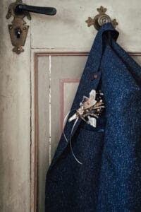 Eine dunkelblaue Jacke mit einer Blumenboutonniere, perfekt für eine Hochzeit, hängt an einer antiken Holztür mit einem verzierten Griff. Die Tür ist in einem verblassten Cremeton mit dezentem rosa Rand gehalten und bietet eine elegante Kulisse für jeden Hochzeitsmoment.
