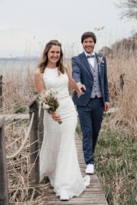 Ein glückliches Paar geht Hand in Hand einen schmalen Holzweg entlang, der von hohen, trockenen Gräsern umgeben ist. Die Braut trägt ein weißes Spitzenkleid und hält einen Blumenstrauß, während der Bräutigam in einem blauen Anzug lächelt. Ihre Hochzeit findet wunderschön im Freien neben einem ruhigen Gewässer statt.