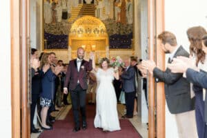 Das Paar, mit der Braut im weißen Kleid und dem Bräutigam im kastanienbraunen Smoking, schreitet bei einer sorgfältig geplanten Hochzeit in einer geschmückten Kirche zum Altar, während auf beiden Seiten die Gäste klatschen.