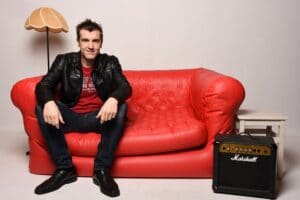 Eine Person in einer schwarzen Lederjacke sitzt auf einem roten Sofa und denkt über ihre Hochzeitsplanung nach. Neben einem kleinen weißen Tisch mit einer Lampe steht ein Marshall-Verstärker auf dem Boden neben dem Sofa und verleiht der Atmosphäre vor dem Hochzeitstag ein Rock'n'Roll-Flair.
