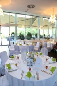 Der Speisesaal ist elegant für eine Hochzeit eingerichtet und verfügt über runde Tische mit weißen Tischdecken. Jeder Tisch ist mit grünen und gelben Servietten, Wasserflaschen, Weingläsern und charmanten Dekorationselementen ausgestattet. Große Fenster bieten einen Blick auf das üppige Grün draußen, perfekt für Ihre Hochzeitsplanung.