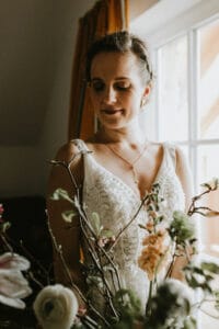 Eine Braut in einem weißen Kleid steht an einem Fenster, das sanft von natürlichem Licht erhellt wird. Während sie ihre Hochzeitsplanung plant, hält sie einen Strauß verschiedener Blumen, darunter Rosen und Blüten, in der Hand und blickt mit einem sanften Lächeln nach unten. Im Hintergrund sind Vorhänge in warmen Farbtönen zu sehen.
