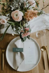 Ein wunderschön gedeckter Tisch für eine Hochzeit, mit einem weißen Teller, einer gefalteten beigefarbenen Serviette und einer Menükarte. Goldfarbenes Besteck ist elegant auf beiden Seiten angeordnet. Das florale Mittelstück, geschmückt mit rosa und weißen Blumen, verleiht Charme, während ein Weinglas die perfekte Hochzeitsplanung abrundet.