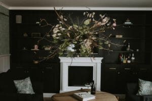 Ein gemütliches Wohnzimmer mit einer schwarzen Wand und eingebauten Regalen, in dessen Mitte ein dekorativer Zweig hängt, der von der Decke hängt – perfekt für die Planung einer Hochzeit. Darunter steht ein runder Holztisch mit einem Buch und einer Weinflasche. Ein weißer Kaminsims und dunkle Möbel runden die Szene ab.