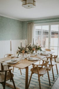 Ein Esszimmer mit einem Holztisch für sechs Personen, perfekt für eine intime Hochzeitsplanung, mit Blumenschmuck und brennenden Kerzen. Holzstühle umgeben den Tisch und Sonnenlicht fällt durch ein Fenster mit beigen Vorhängen. Die Wände sind in einem gedämpften Grün mit weißer Wandvertäfelung gehalten.