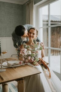 Ein Paar posiert an einem großen Fenster und verkörpert die Essenz einer Hochzeit. Der Mann im Anzug küsst sanft die Schläfe der Frau, während sie lächelt und einen Strauß rosa und weißer Blumen in der Hand hält. Neben ihnen verstärkt ein gedeckter Esstisch die ruhige Atmosphäre, perfekt für ihre Hochzeitsfeier.