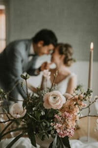 Ein Paar lehnt sich an einem sanft beleuchteten, mit Blumen geschmückten Tisch, darunter rosa Rosen und Ranunkeln, zu einem Kuss vor und fängt damit die Essenz einer romantischen Hochzeit perfekt ein. Eine brennende Kerze verleiht der gemütlichen Atmosphäre Wärme und schafft eine ideale Bühne für Liebe und intime Hochzeitsplanung.