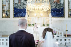 Ein Paar steht vor einem Priester in einer Kirche und blickt auf einen reich verzierten Altar mit einem goldenen Baldachin, der ihre sorgfältige Hochzeitsplanung widerspiegelt. Der Innenraum ist mit aufwendigen Mustern dekoriert und im Vordergrund ist ein Blumenarrangement zu sehen.