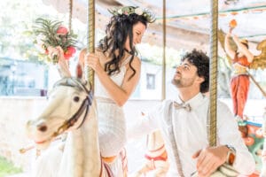 Braut und Bräutigam auf einem Karussell lächeln sich an, während sie ihre Hochzeit feiern. Die Braut hält einen Blumenstrauß und trägt eine Blumenkrone, während der Bräutigam, in ein weißes Hemd und eine Fliege gekleidet, neben ihr sitzt. Helle und fröhliche Kulisse mit einer Engelsfigur im Hintergrund.