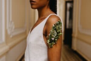 In einer Szene, die an elegante Hochzeitsplanung erinnert, steht eine Person mit geflochtenen Haaren in einem Flur mit verzierten weißen Wänden und gemustertem Boden und trägt ein weißes ärmelloses Oberteil. Ein detailreiches Blumenarrangement schlingt sich elegant um ihren Oberarm und verleiht dieser von einer Hochzeit inspirierten Szene Charme.