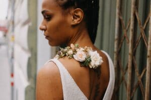 Eine Frau mit geflochtenem Haar und einem ärmellosen Oberteil steht mit dem Rücken zu ihr. Auf ihrer linken Schulter ruht ein Blumenarrangement aus weißen und hellrosa Rosen, was einen Hauch von Hochzeitseleganz suggeriert. Im Hintergrund sind eine Metallstruktur und eine verschwommene Straßenansicht zu sehen.