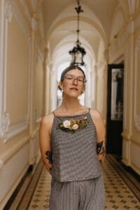 Eine Person mit Brille und Tattoos steht in einem Flur mit verzierten Zierleisten und verkörpert einen einzigartigen Stilmix, der perfekt zu einer Hochzeit passt. Sie trägt ein kariertes, mit Blumen geschmücktes Oberteil und eine passende Hose. Eine Hängelampe und eine gewölbte Decke vervollständigen die elegante Kulisse.