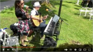 Eine Frau singt mit einem Mikrofon, während ein Mann Gitarre spielt und neben einem Keyboard im Freien auf einer Rasenfläche sitzt, perfekt für eine Hochzeit. Weiße Stühle und Bäume im Hintergrund verstärken die lockere und lebhafte Atmosphäre, ideal für unvergessliche Hochzeitsplanungsmomente.