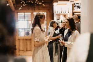 Zwei Frauen unterhalten sich in einem warm beleuchteten Raum, jede hält ein Glas Champagner in der Hand. Eine trägt ein weißes Kleid mit Spitzenärmeln – eine Anspielung auf die Eleganz einer Hochzeit – und die andere ist dunkel gekleidet und trägt einen weißen Mantel. Menschen und Holzdekor im Hintergrund lassen subtile Andeutungen von Hochzeitsplanung vermuten.