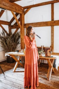 Eine Frau in einem langen orangefarbenen Kleid spricht in ein Mikrofon in einem rustikalen Raum mit Holzbalken und bespricht wahrscheinlich die Hochzeitsplanung. Hinter ihr steht ein Holztisch, der mit einem dekorativen Buch und getrockneten Pflanzen geschmückt ist und das perfekte Ambiente zum Heiraten schafft.