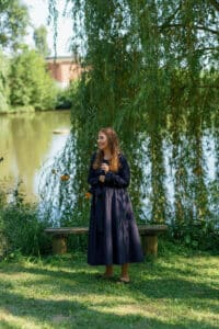 Eine Person in einem dunklen Kleid steht auf Gras vor einem Teich, eingerahmt von herabhängenden Weidenblättern – ein ruhiger Ort, perfekt für Hochzeitsfotos. Sie scheint sich an einem sonnigen Tag in einem Park zu befinden, mit einer Bank in der Nähe und einem Hintergrund voller Grün und ruhigem Wasser.