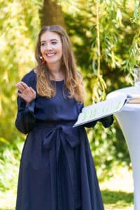 Eine Frau mit langen braunen Haaren und einem dunkelblauen Kleid lächelt und winkt, während sie ein Dokument in der Hand hält. Sie steht im Freien neben einem hohen Tisch mit üppigem Grün im Hintergrund und verkörpert perfekt die Freude der Hochzeitsplanung.