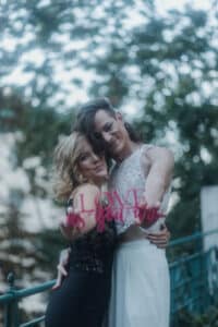 Zwei Menschen stehen dicht beieinander im Freien, lächeln und halten ein Schild mit der Aufschrift „LIEBE, wie du bist“. Die eine trägt ein schwarzes Kleid und die andere ein weißes Spitzentop mit weißen Hosen. Sie stehen auf einer Brücke mit Bäumen im Hintergrund – ein perfekter Moment der Liebe, festgehalten während ihrer Hochzeitsreise.