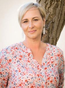Eine Frau mit kurzen blonden Haaren steht vor einem Baum und strahlt Freude an ihrem Hochzeitstag aus. Sie trägt eine Bluse mit Blumenmuster und weiße Ohrringe, lächelt sanft in die Kamera und denkt vielleicht über die nächsten Schritte ihrer Hochzeitsplanung nach.