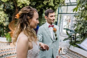 Während der Hochzeit stehen Braut und Bräutigam in einer üppigen, grünen Umgebung zusammen. Die Braut trägt ein weißes Kleid, während der Bräutigam einen hellgrünen Anzug mit einer roten Fliege und einem Blumenboutonniere trägt. Kerzen und Blätter rahmen diese bezaubernde Hochzeitsszene ein.